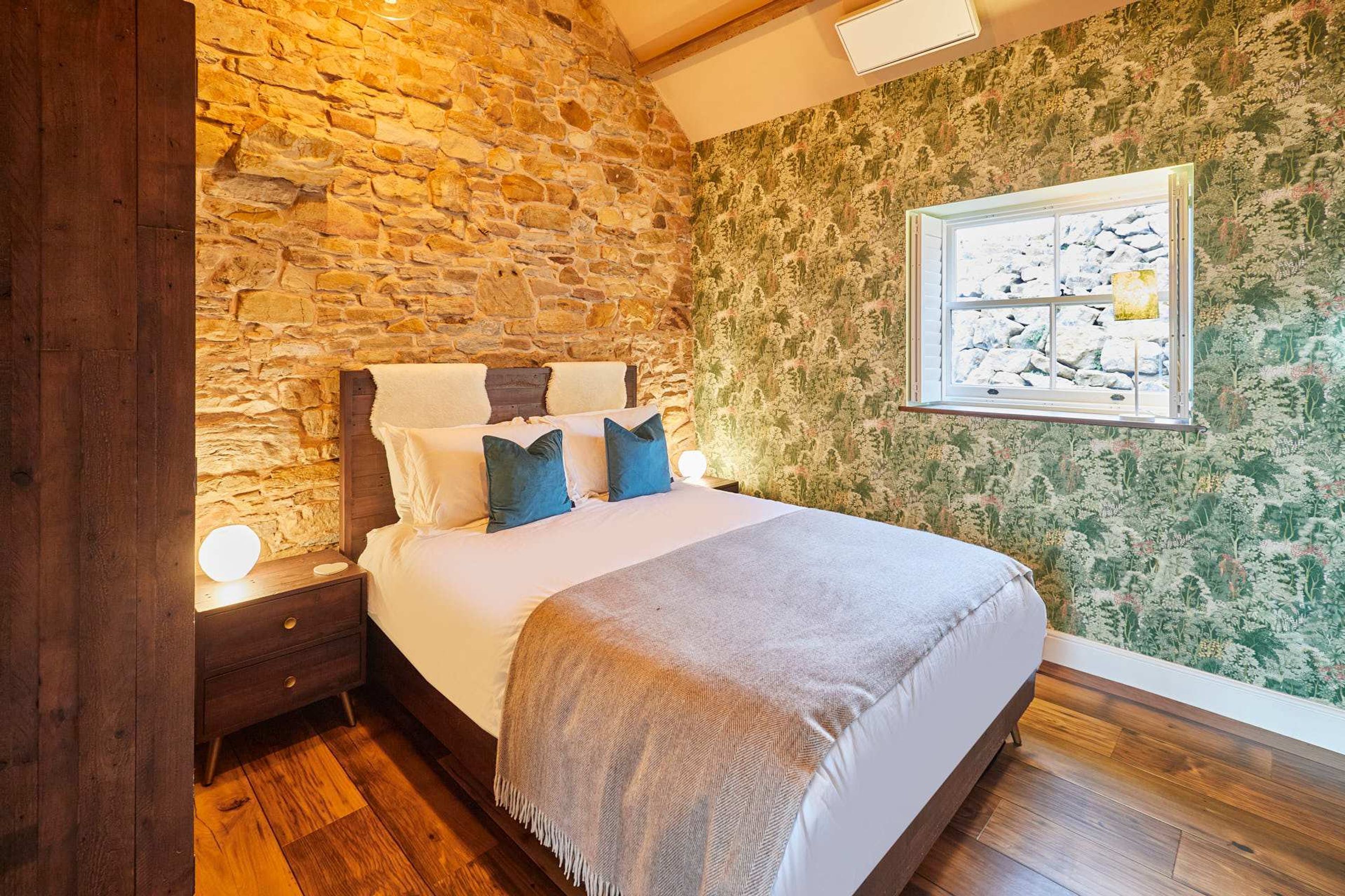 Une chambre avec un mur en pierre, un papier peint à motifs floraux, un lit avec des oreillers, une lampe de chevet et une fenêtre donnant sur un paysage agréable.