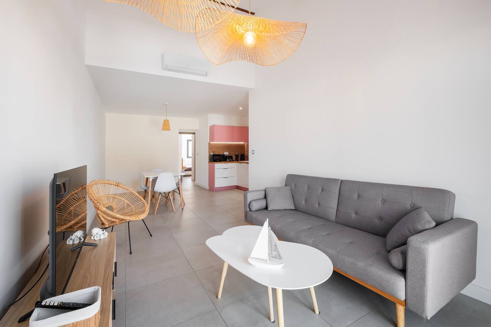 A modern living room with a grey sofa, white coffee table, wicker chairs, and a TV. A kitchen with pink cabinets is visible in the background.