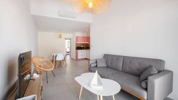 A modern living room with a grey sofa, white coffee table, wicker chairs, and a TV. A kitchen with pink cabinets is visible in the background.