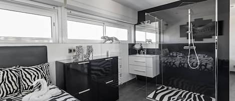 A modern hotel room with a zebra-patterned bed, a black wardrobe, a white vanity, a shower with a glass enclosure, and a large window providing natural light.