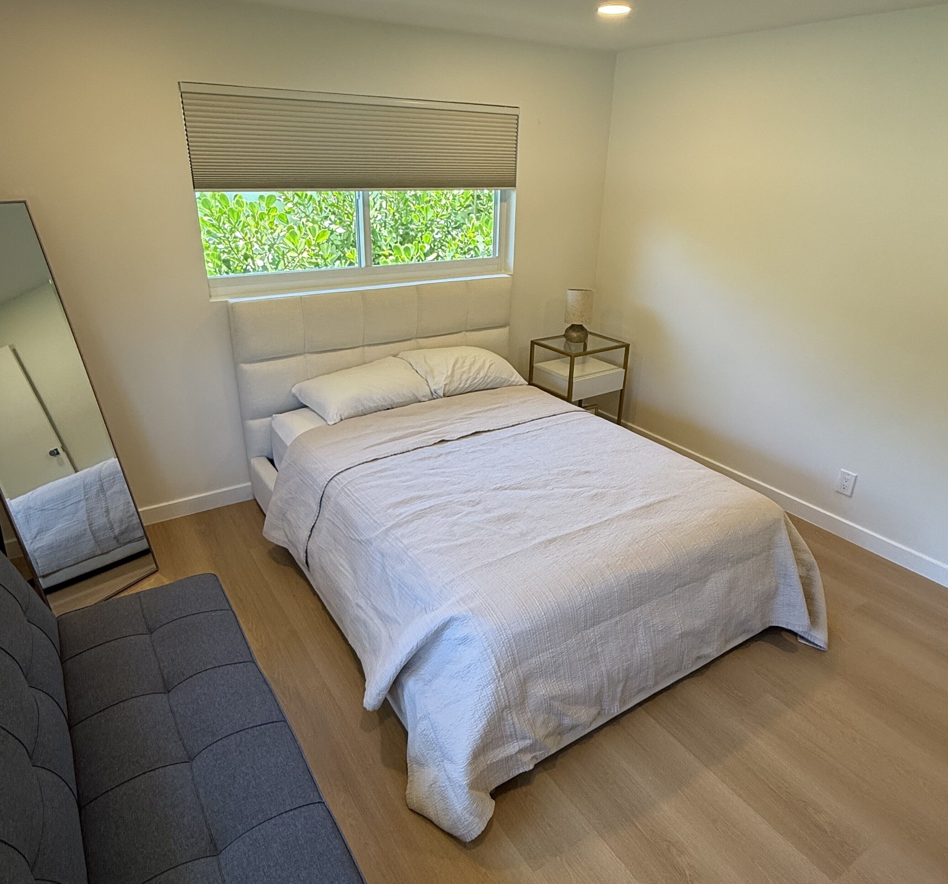 Ein Schlafzimmer mit einem Bett, einem Nachttisch, einem Fenster mit Jalousien, einem Spiegel und einem grauen Sofa.
