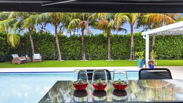 Zona de piscina con mesa, sillas y vistas a palmeras y seto.
