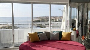 En rund seng med rødt lagen, hvid base og mange puder i et rum med store vinduer med udsigt over strand og bybillede.