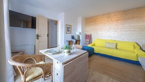A compact living space with a kitchenette, a dining area with wicker furniture, a bright yellow sofa, and a wooden wall. - Le Nid de Cabourg - Welkeys (Cabourg)
