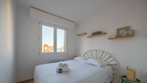 A neatly made bed with rolled towels, a bedside table with a basket, and a window with a view of buildings. - Le Nid de Cabourg - Welkeys (Cabourg)