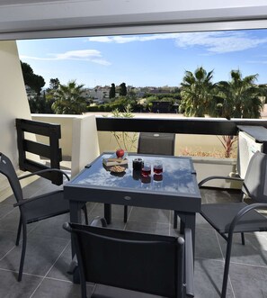 A glass-top table with chairs, a bowl of nuts, a book, and two glasses of red wine on a balcony with a view of palm trees and houses. - GENTIANE - Apartment Port Nature 475 - Luxury - 2 people (AGDE)