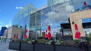 Een modern gebouw met grote glasramen, een bord met 'ERIN MILLS TOWN CENTER' en terrasmeubilair met rode parasols.