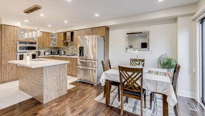 Een moderne keuken met een centraal eiland, roestvrijstalen apparatuur, houten kastjes en een eethoek met een tafel en stoelen.