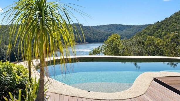 A clear swimming pool with a wooden deck, surrounded by lush greenery and a view of a lake and hills.