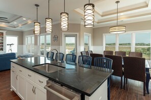 A kitchen with a large island, a dining area with chairs, and a view of the outdoors. - Why Knot | Pool, Elevator, Putting Green, Cabana Service & $250 Beach Gear Credit – Across from Beach Access (Holden Beach)