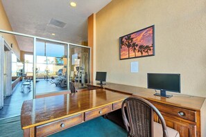 Ein Hotelzimmer mit einem Holztisch, zwei Monitoren, einem Korbstuhl und einer Glaswand, die in einen Fitnessbereich führt.