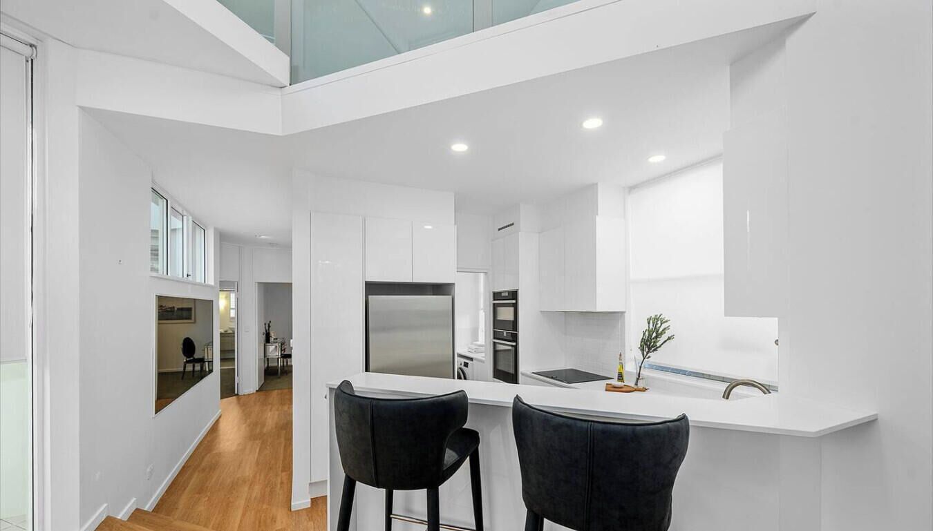 A modern kitchen with white cabinetry, a stainless steel refrigerator, and a bar area with two black chairs.