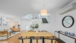 Ein modernes Wohnzimmer mit Esstisch aus Holz, Korbstühlen, einer Treppe und einem großen Spiegel an der Wand.