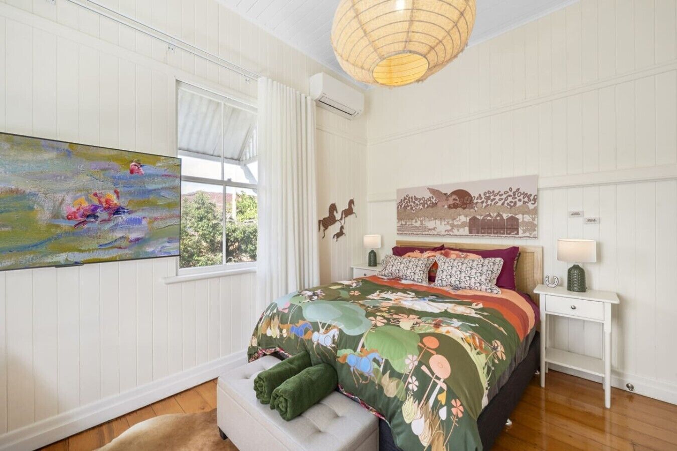 A bedroom with a large bed, a window with curtains, a painting on the wall, and a bedside table with a lamp.