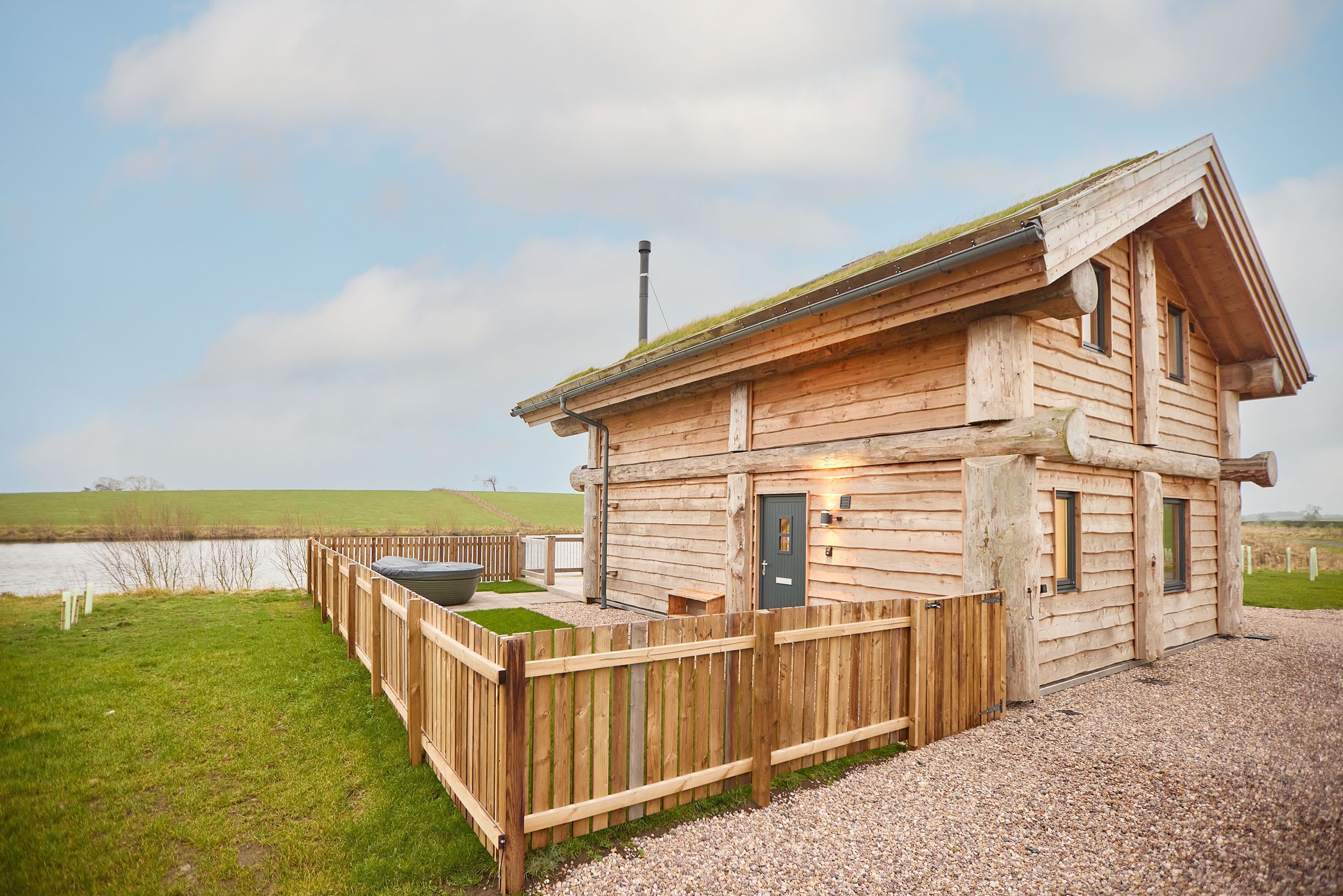 Une cabane en bois avec une allée en gravier, une cour clôturée et un plan d’eau en arrière-plan.