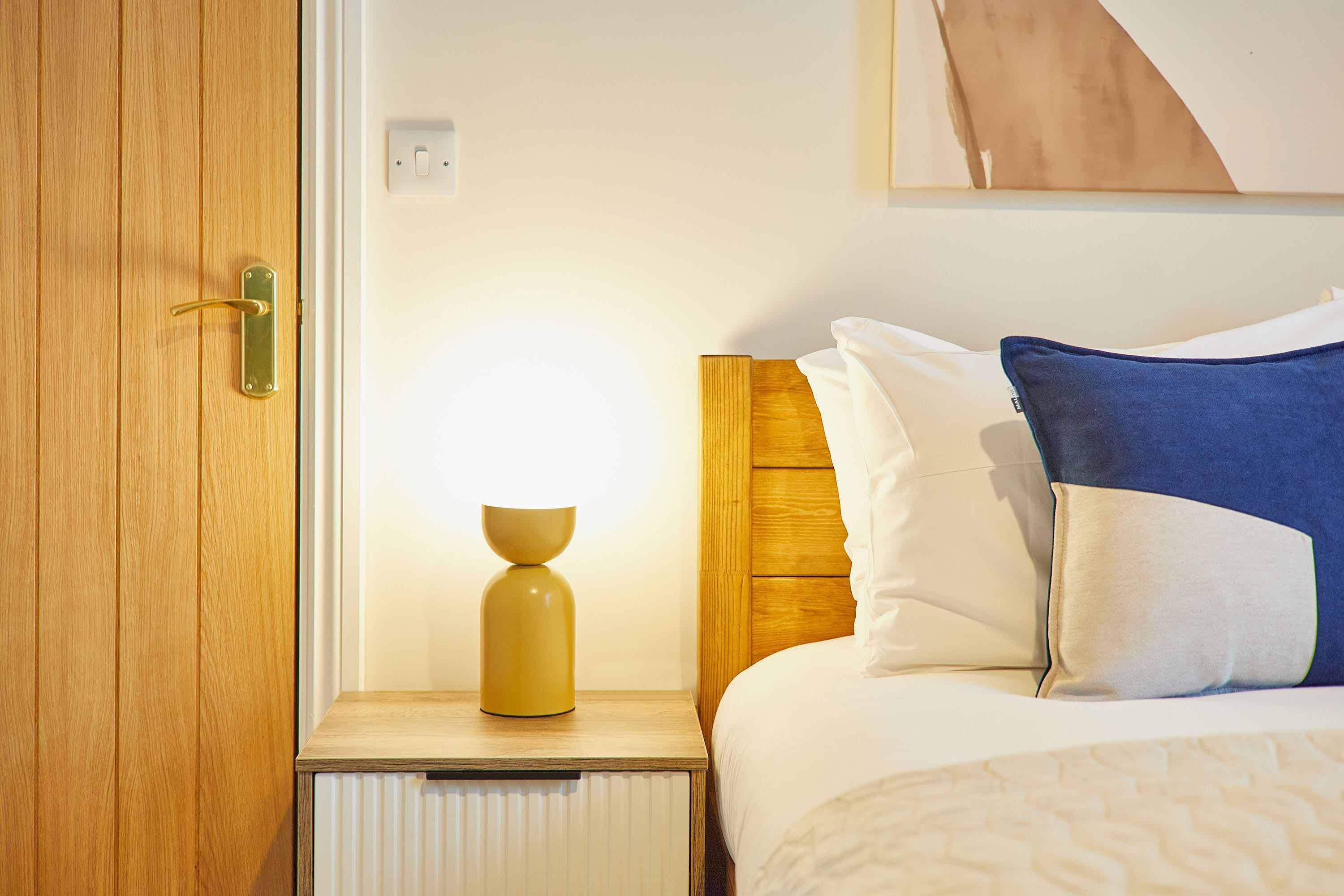 A wooden door with a brass handle, a bedside table with a lamp, and a bed with white and blue pillows.