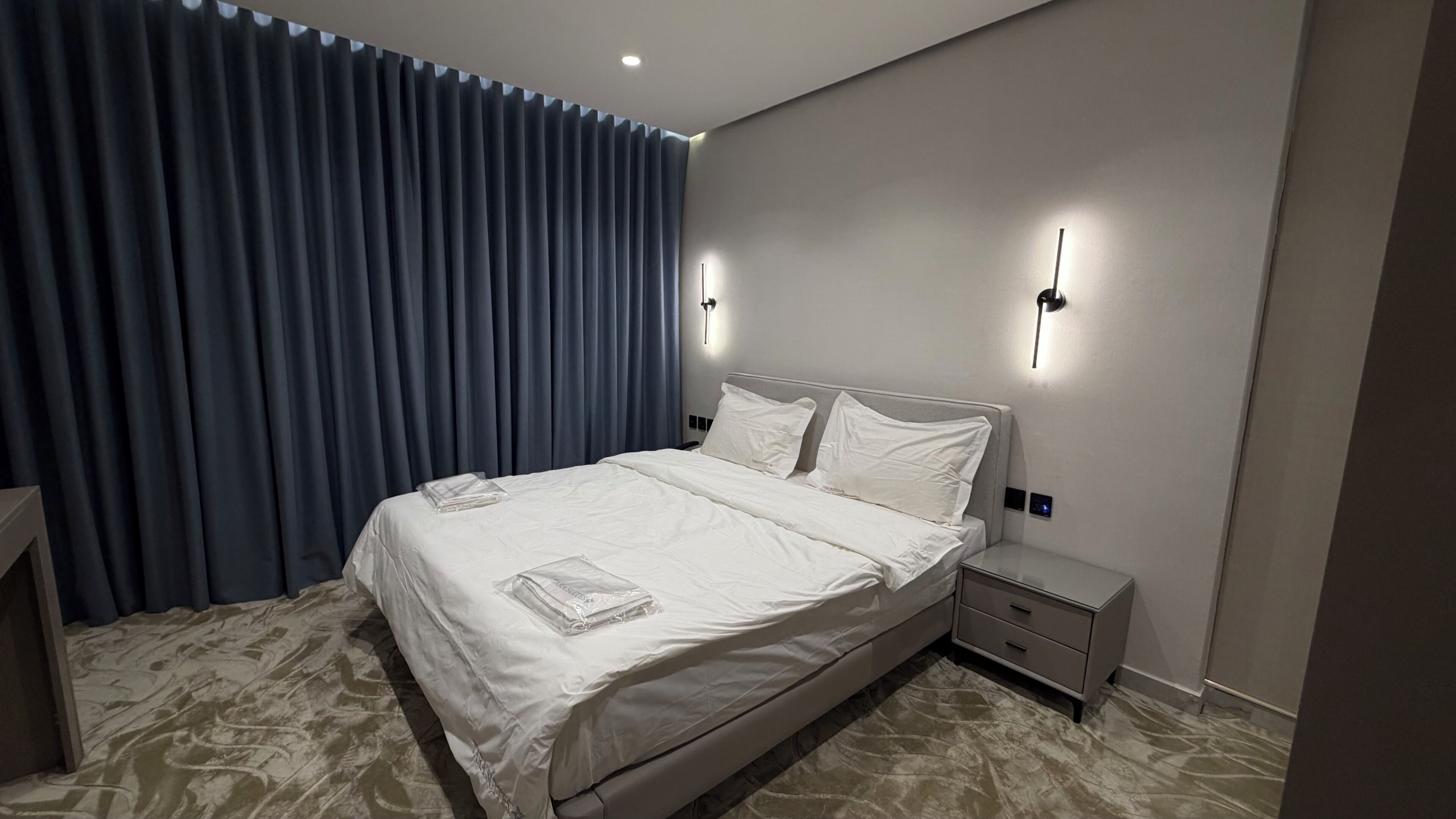 A hotel room with a large bed, bedside tables, a nightstand, and a wall-mounted light fixture.