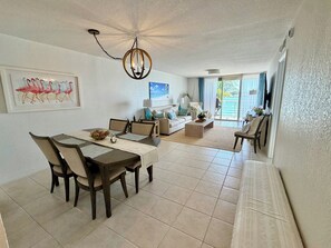 A spacious living area with a dining table, chairs, a sofa, and a coffee table. There is a ceiling light fixture, a painting of flamingos, and a view through a glass door to the outside. - Waterfront 2BR, 2BA condo. Newly updated St. Pete, St. Petersburg, Isla Del Sol (St. Petersburg)