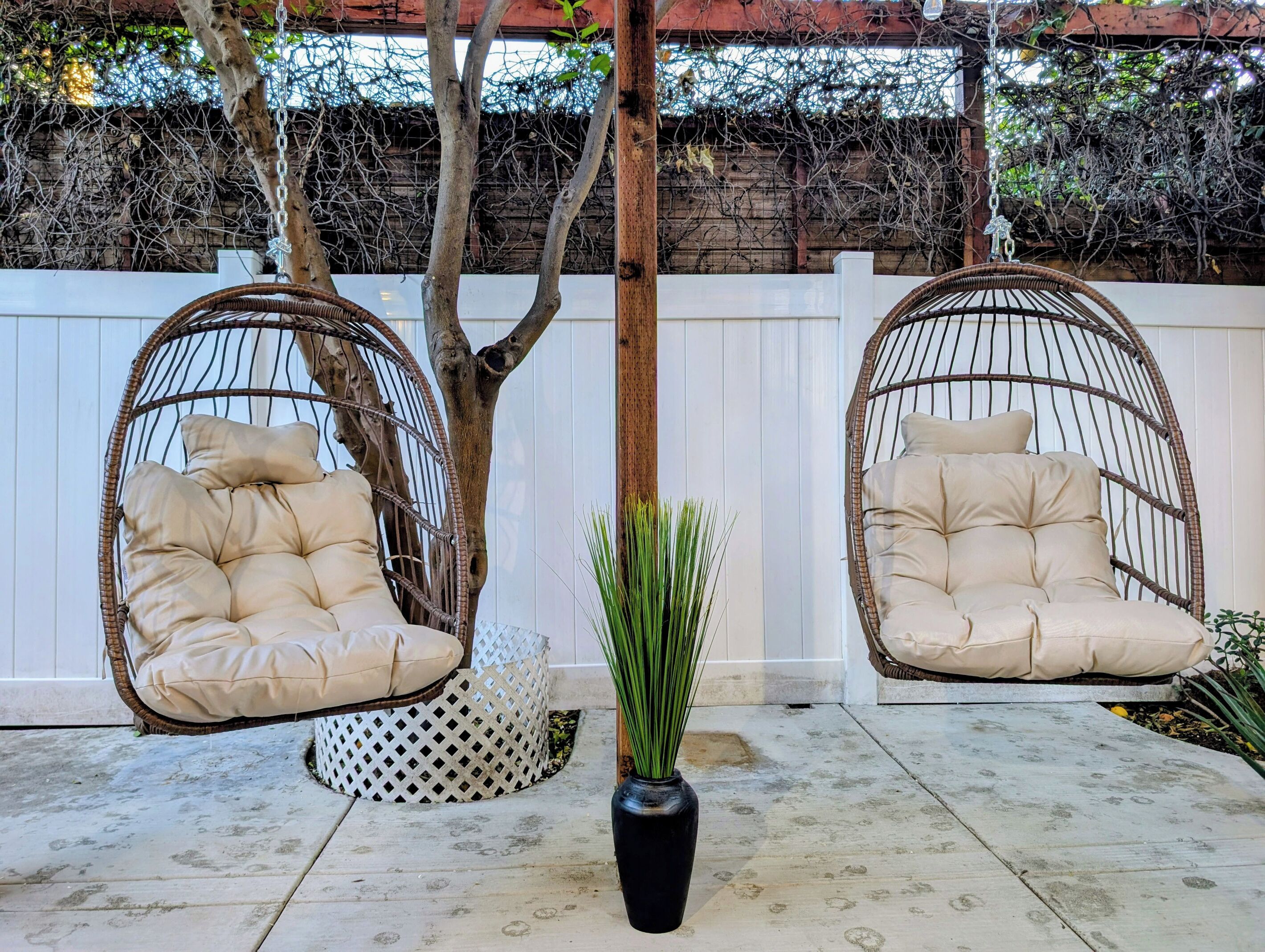 Two wicker swing chairs with cushioned seats and backrests, positioned on a concrete patio with a white fence and trees in the background.