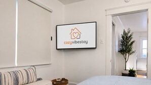 Een slaapkamer met een bed, een bank, een aan de muur bevestigde televisie waarop een logo staat, en een hal met een plant.