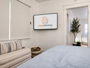 A bedroom with a bed, a sofa, a wall-mounted TV displaying a logo, and a hallway with a plant. - CozyVibeStay. Walk to Sand. Workspace. Parking (Long Beach)
