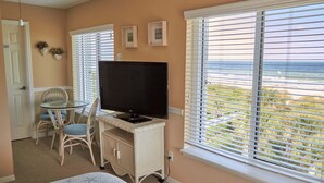 Ein Schlafzimmer mit Bett, Fernseher auf einer weißen Kommode, Esstisch mit Stühlen und einem Fenster mit Jalousien.
