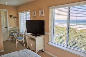 Ein Schlafzimmer mit Bett, Fernseher auf einer weißen Kommode, Esstisch mit Stühlen und einem Fenster mit Jalousien.