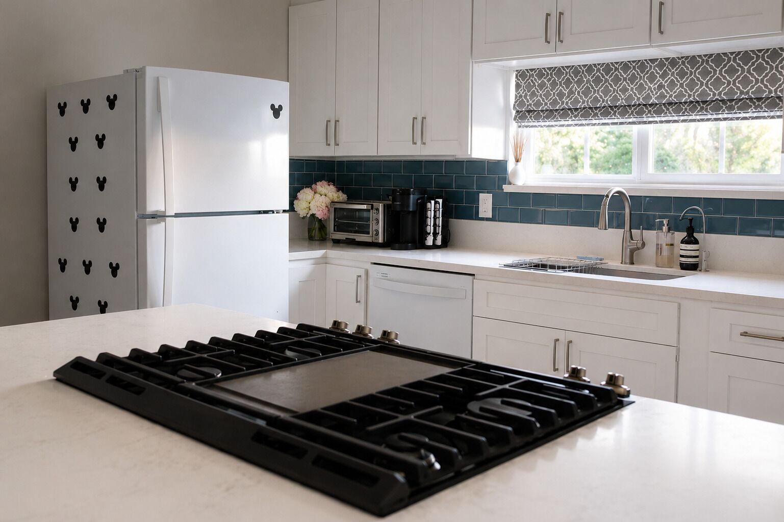 A modern kitchen with white cabinets, a built-in stove, a refrigerator with Mickey Mouse magnets, a toaster, and a window with a patterned valance.
