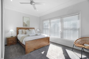 A bedroom with a bed, bedside tables, a ceiling fan, a window with blinds, and a wicker chair.
