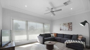 A modern living room with a sectional sofa, a round coffee table, a ceiling fan, and a large window with blinds.