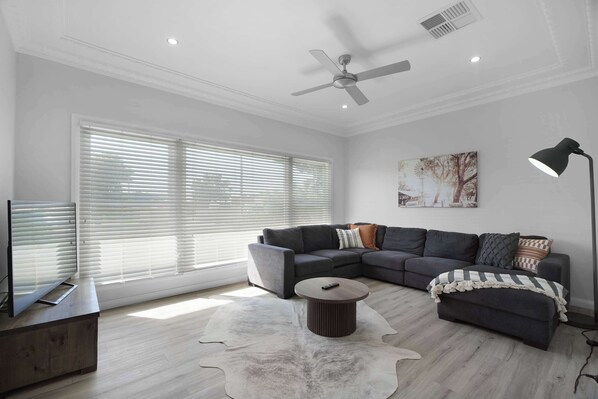 A modern living room with a sectional sofa, a round coffee table, a ceiling fan, and a large window with blinds.