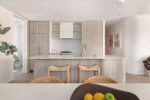 A modern kitchen with a central island, wooden bar stools, and a dining table. - 3BR Coastal Apartment - Augusta 203 (Burleigh Heads)
