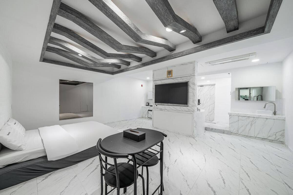 A modern hotel room with a bed, a small table, a TV, and a bathroom with a mirror and sink.