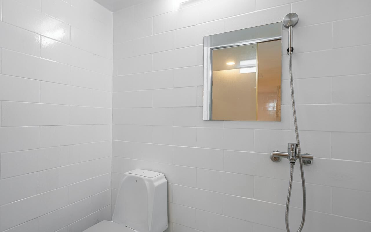 Un baño con paredes de azulejos blancos, un cabezal de ducha de mano, un inodoro y un espejo.