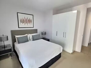 Quarto moderno com uma cama grande, mesa de cabeceira com luminária, guarda-roupa e uma obra de arte emoldurada na parede.