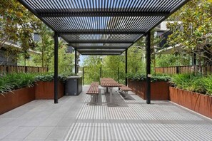 Um pátio externo com mesa e bancos de madeira, cercado por vasos de plantas e uma estrutura de pérgola de metal.