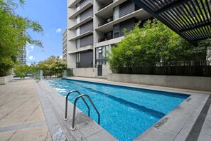 Piscina com escada de metal, cercada por um deck de concreto e vegetação.