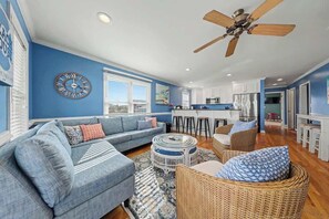 A living room with a sofa, a coffee table, and a wicker chair. There is a ceiling fan, a kitchen area with a refrigerator, and a dining table. - Sun Sprinkles by Carolina Getaway - Ocean Views, Pool, Walk to Kure Beach (Kure Beach)