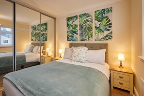 A hotel room with a bed, a nightstand, a lamp, a mirror, and tropical leaf artwork on the wall. - Host Stay Bridge Court (Yarm)