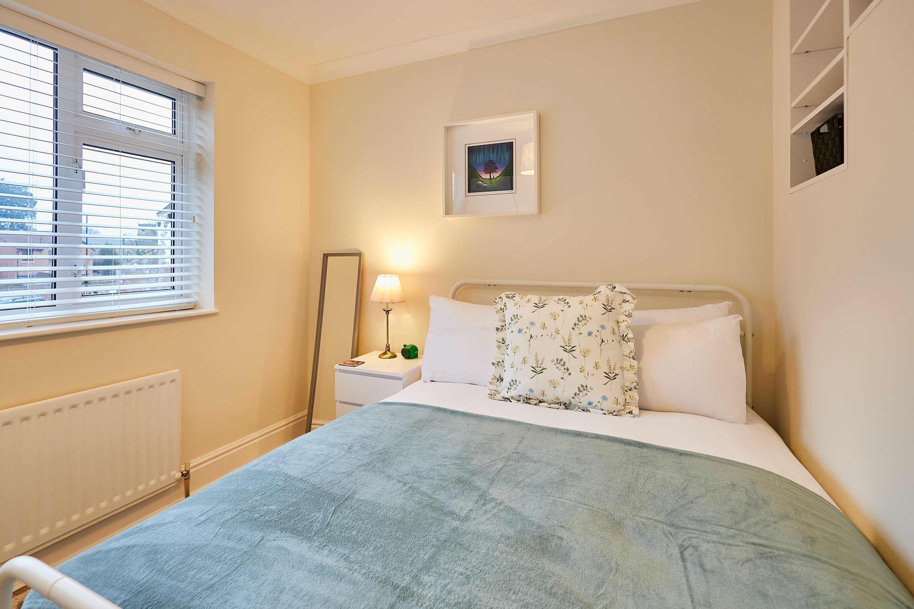 A bedroom with a bed, a nightstand, a lamp, a window with blinds, a radiator, and a framed picture on the wall.