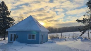 Uma pequena casa azul com telhado empena, rodeada por neve e árvores, sob um céu parcialmente nublado.