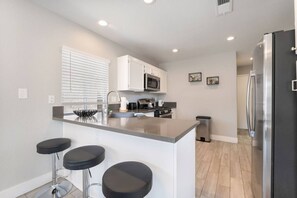A modern kitchen with white cabinets, a stainless steel refrigerator, a microwave, and a bar stool area. - Modern 3BR Home | Pets+7 Min to Airport+Parking (Las Vegas)