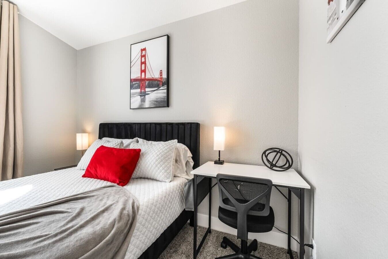 Habitación de hotel con cama, escritorio, silla, lámpara y un cuadro del Puente Golden Gate.