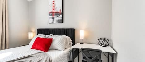 A hotel room with a bed, a desk, a chair, a lamp, and a picture of the Golden Gate Bridge.