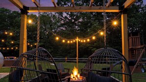 Un espacio al aire libre techado con dos sillones colgantes, una chimenea y luces decorativas.
