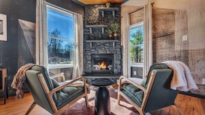 Una sala de estar acogedora con chimenea de piedra, dos sillones verdes, una mesa redonda de centro y ventanas amplias.
