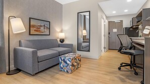 Ein modernes Hotelzimmer mit grauem Sofa, einem gemusterten Hocker, einem großen Spiegel, einem Schreibtisch mit Stuhl und einer Küchenzeile.