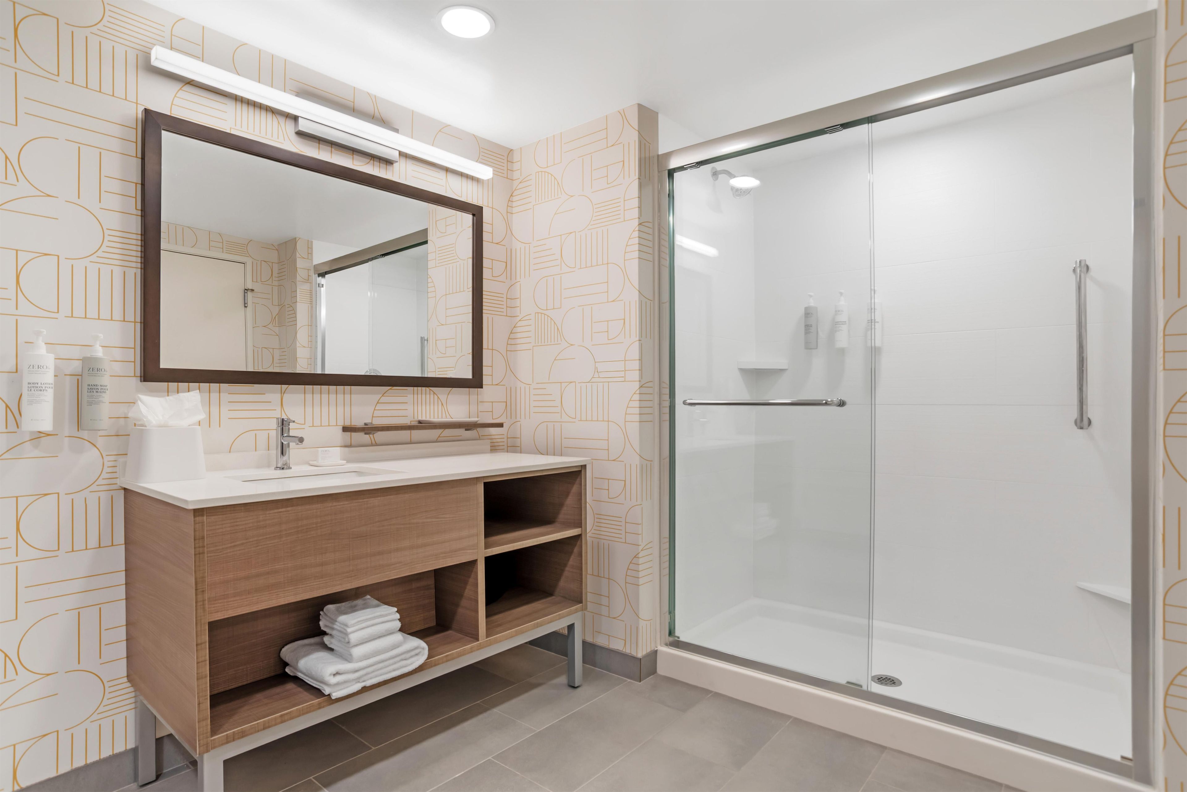 A modern bathroom with a glass shower enclosure, a wooden vanity with a mirror, and patterned wall tiles.