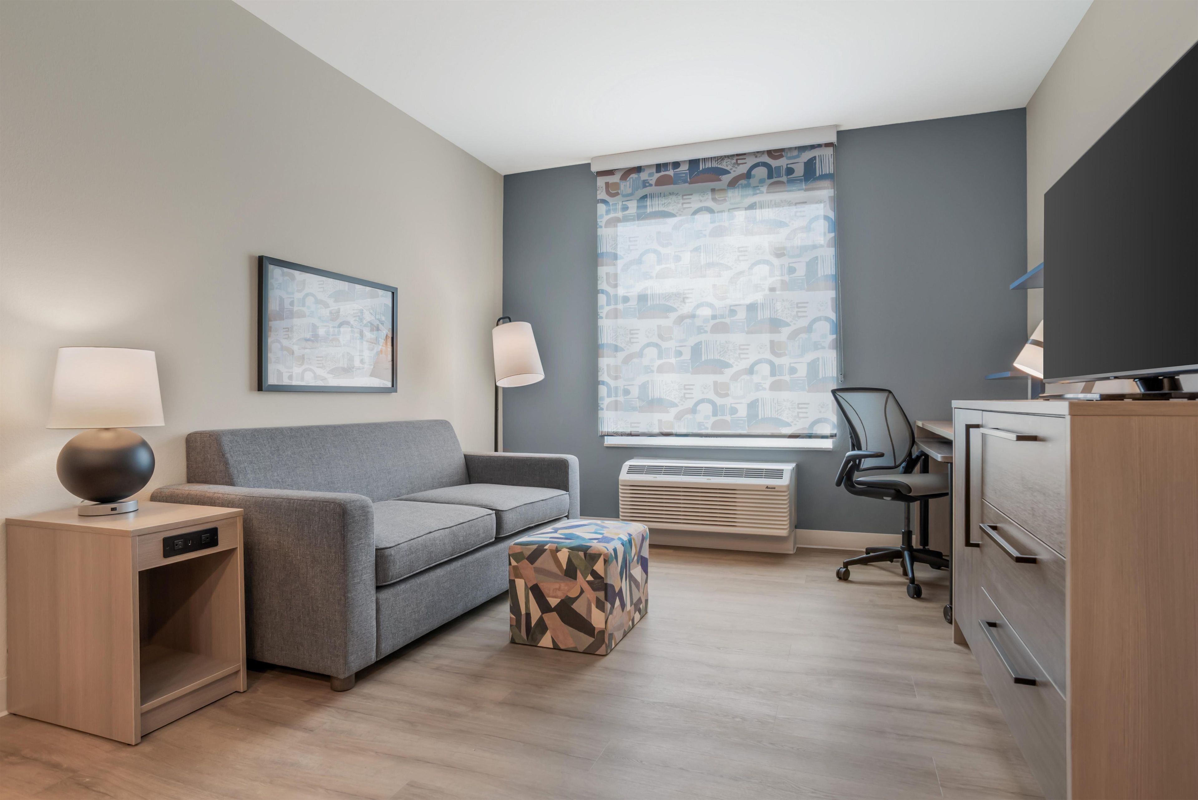 Une chambre d’hôtel moderne avec un canapé gris, une table d’appoint en bois avec une lampe, un pouf à motifs, un bureau avec une chaise et une grande fenêtre ornée de rideaux à motifs.