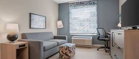 Une chambre d’hôtel moderne avec un canapé gris, une table d’appoint en bois avec une lampe, un pouf à motifs, un bureau avec une chaise et une grande fenêtre ornée de rideaux à motifs.